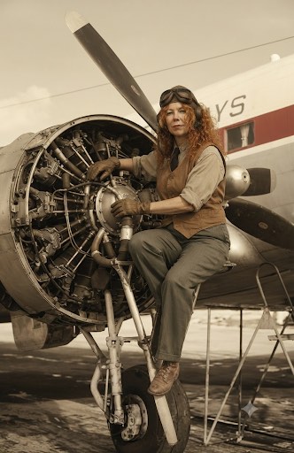 A vintage sepia-toned photograph of a woman in pilot gear sitting on the landing gear of a propeller plane.